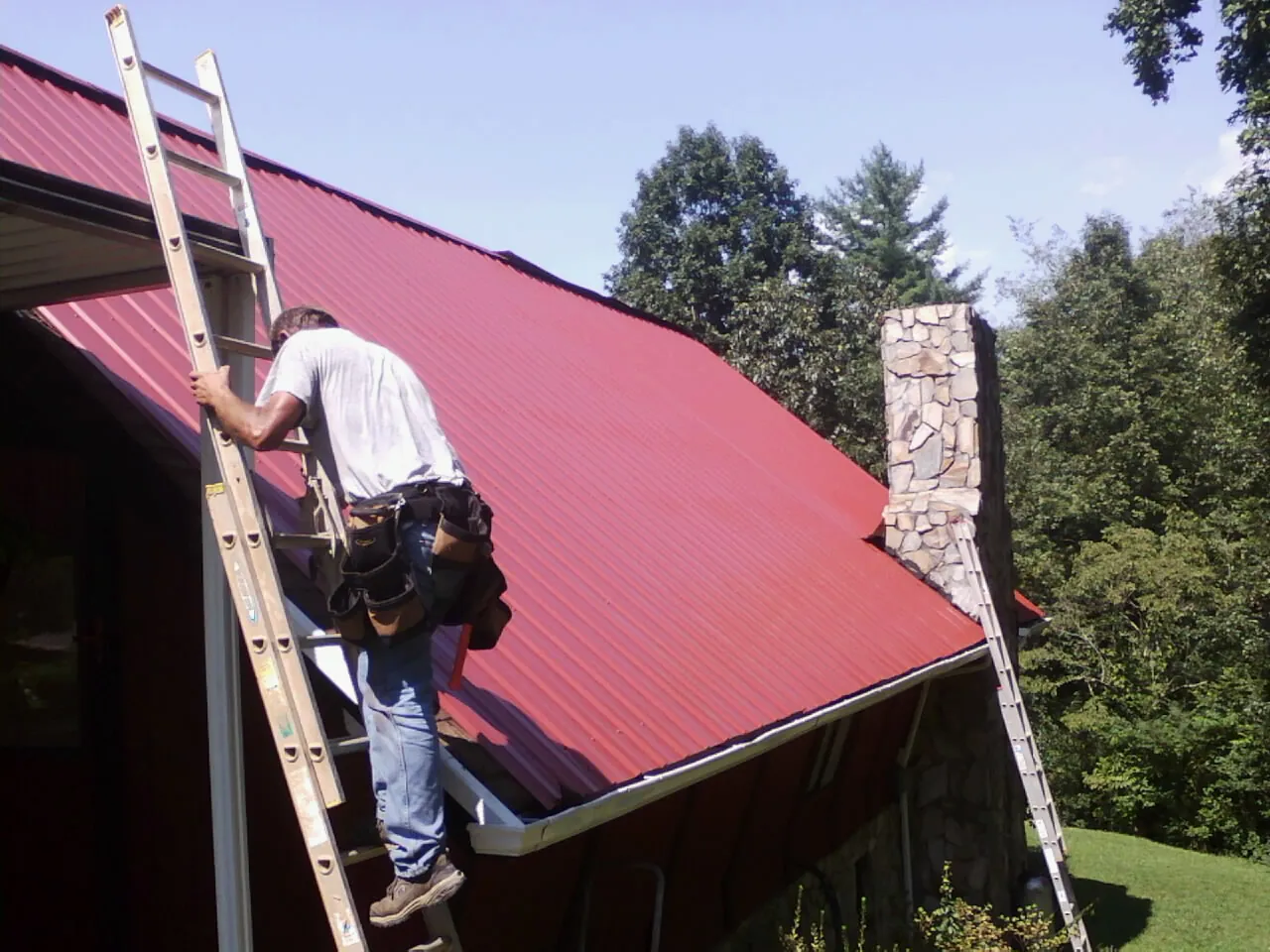 Roofer on a ladder beside a red metal roof and stone chimney — Soffit & Fascia Repair by Roofing Brothers in McCall